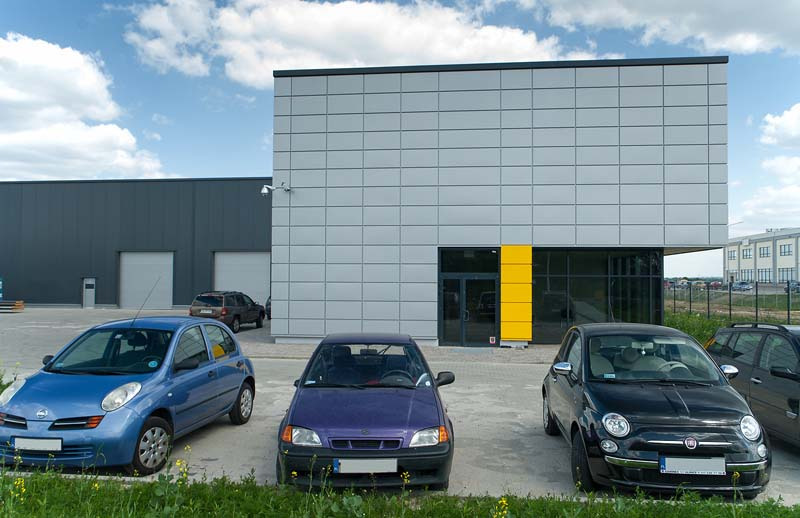 Nowoczesny budynek przemysłowy z szarej płyty warstwowej, z żółtym akcentem przy wejściu, widok z parkingu z trzema zaparkowanymi samochodami w różnych kolorach.