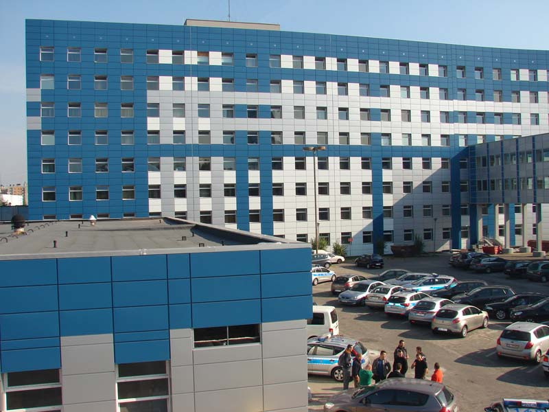 Komenda Wojewódzka Policji w Katowicach: budynek obłożony niebiesko-szarymi panelami warstwowymi, widok z góry na parking pełen samochodów.