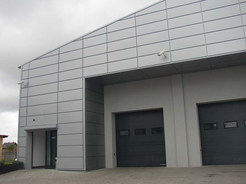 Szary budynek przemysłowy z elewacją z paneli, widoczne dwie bramy garażowe i wejście boczne, kamera monitoringu na ścianie, pochmurne niebo.