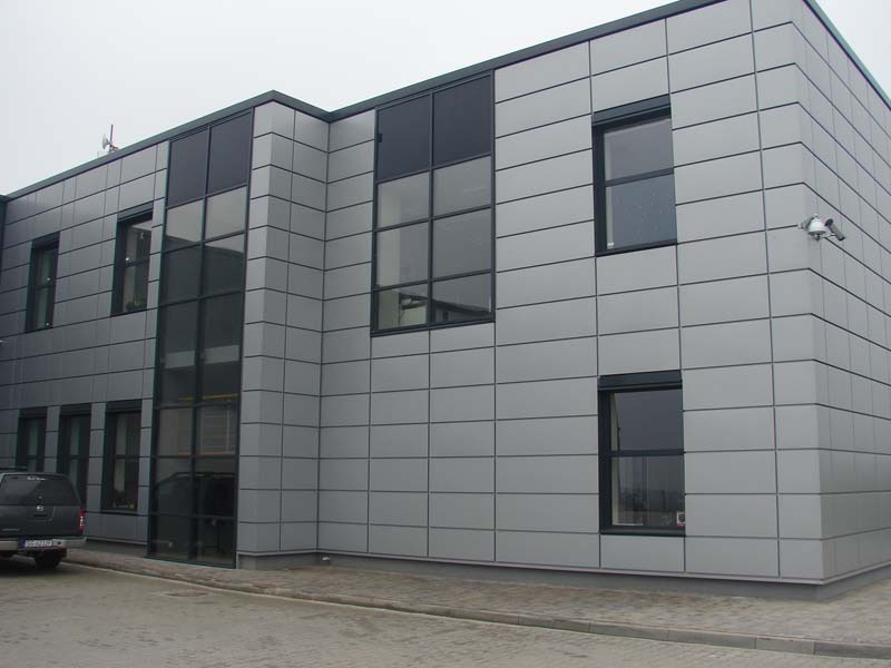 Budynek biurowy wykończony panelami elewacyjnymi w odcieniach szarości, z widocznym fragmentem parkingu i samochodu osobowego w lewym dolnym rogu.