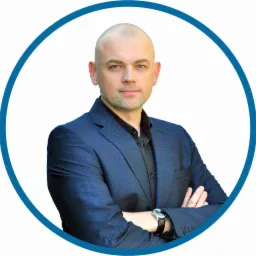 Portret mężczyzny w niebieskiej marynarce, z zegarkiem na ręce, na białym tle w niebieskim okręgu, z założonymi rękami i lekkim uśmiechem.