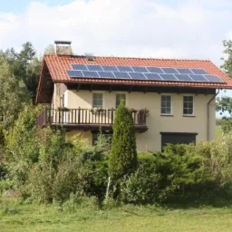 Dwupiętrowy dom z beżową elewacją i czerwonym dachem, na którym zamontowane są panele słoneczne. Balkon z drewnianą balustradą ozdobiony kwiatami. Dom otoczony zielenią.