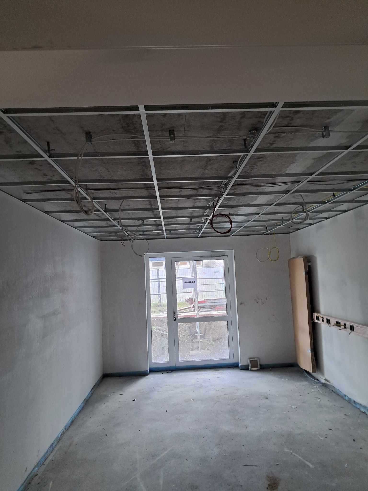 Montaż metalowego rusztu podwieszanego sufitu w surowym pomieszczeniu z widocznymi przewodami elektrycznymi i przygotowanymi ścianami.