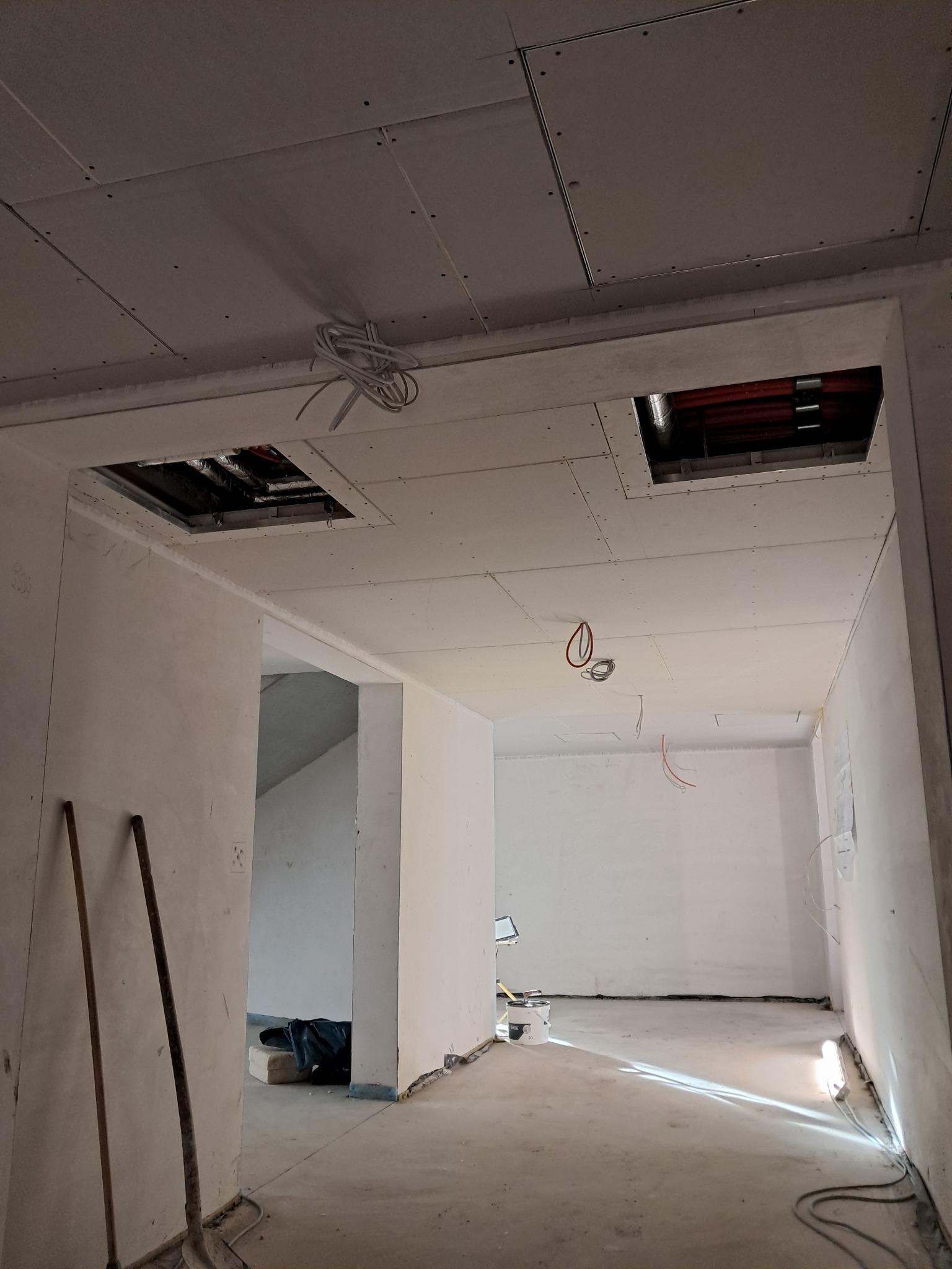 Wnętrze w trakcie montażu sufitu podwieszanego z płyt kartonowo-gipsowych, widoczne otwory rewizyjne z instalacjami, przewody elektryczne i narzędzia malarskie oparta o ścianę.