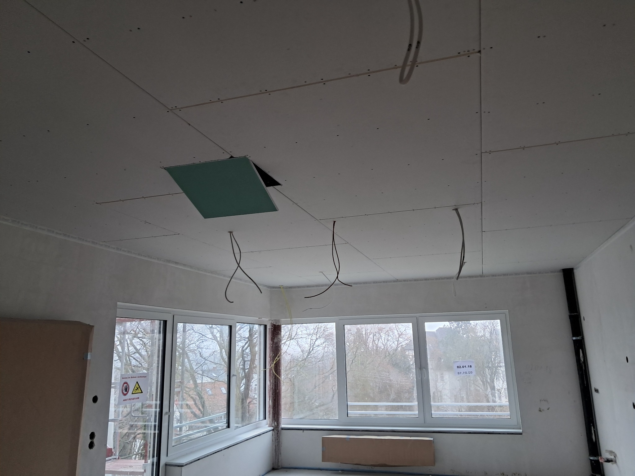 Sufit z płyt kartonowo-gipsowych w trakcie montażu, widoczne kable, otwór rewizyjny i okna z widokiem na drzewa.