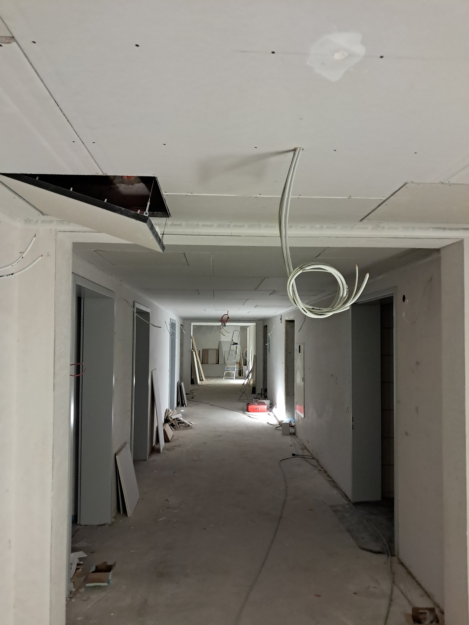 Korytarz w trakcie prac wykończeniowych: widoczne płyty karton gips na suficie z otworami, zwisające kable elektryczne, przygotowane ściany przed malowaniem.