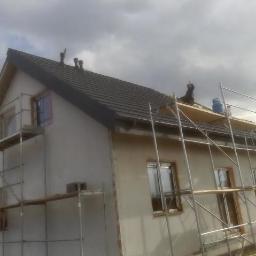 MARL - Dwukondygnacyjny dom w trakcie budowy, z widocznym rusztowaniem i pracownikiem na dachu, obok butla z gazem, pochmurne niebo w tle.