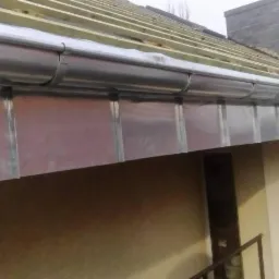 Nowe, metalowe rynny zamontowane na budynku z widoczną konstrukcją dachu i kominem w tle, wykonane w pochmurny dzień.