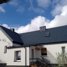 Biały dom jednorodzinny z ciemnoszarym dachem w kształcie dachówki, antena telewizyjna na ścianie, błękitne niebo z chmurami w tle.