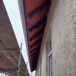 MARL - Widok z dołu na okap dachu z drewnianą podbitką i metalową rynną, obok rusztowanie. Ściana budynku o szorstkiej fakturze.