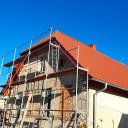 MARL - Budynek w trakcie remontu dachu, widoczne rusztowanie, nowa czerwona dachówka i fragmenty starego muru z cegły.