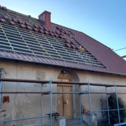 Częściowo ułożona dachówka ceramiczna na łatach na dachu starego domu z szarymi ścianami i drewnianymi drzwiami, widoczne rusztowanie.