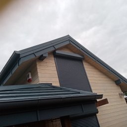 MARL - Fragment nowoczesnego domu z drewnianą elewacją, ciemnoszarym dachem z blachy i roletami zewnętrznymi, widoczny z dołu pod kątem, pochmurne niebo w tle.