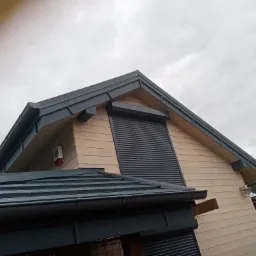 Fragment nowoczesnego domu z drewnianą elewacją, ciemnoszarym dachem z blachy i roletami zewnętrznymi, widoczny z dołu pod kątem, pochmurne niebo w tle.