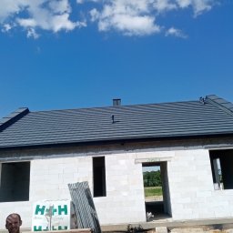 MARL - Surowy stan domu z białego bloczka z zamontowanym szarym dachem pod błękitnym niebem z chmurami, widoczne materiały budowlane i uśmiechnięta osoba w rogu kadru.