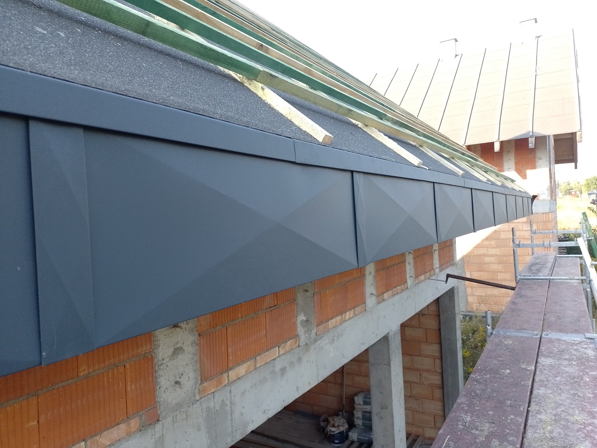 Szary panel okapowy z geometrycznym wzorem na nowo budowanym domu w Lwówku, widok z rusztowania na fragment dachu z widoczną konstrukcją drewnianą.