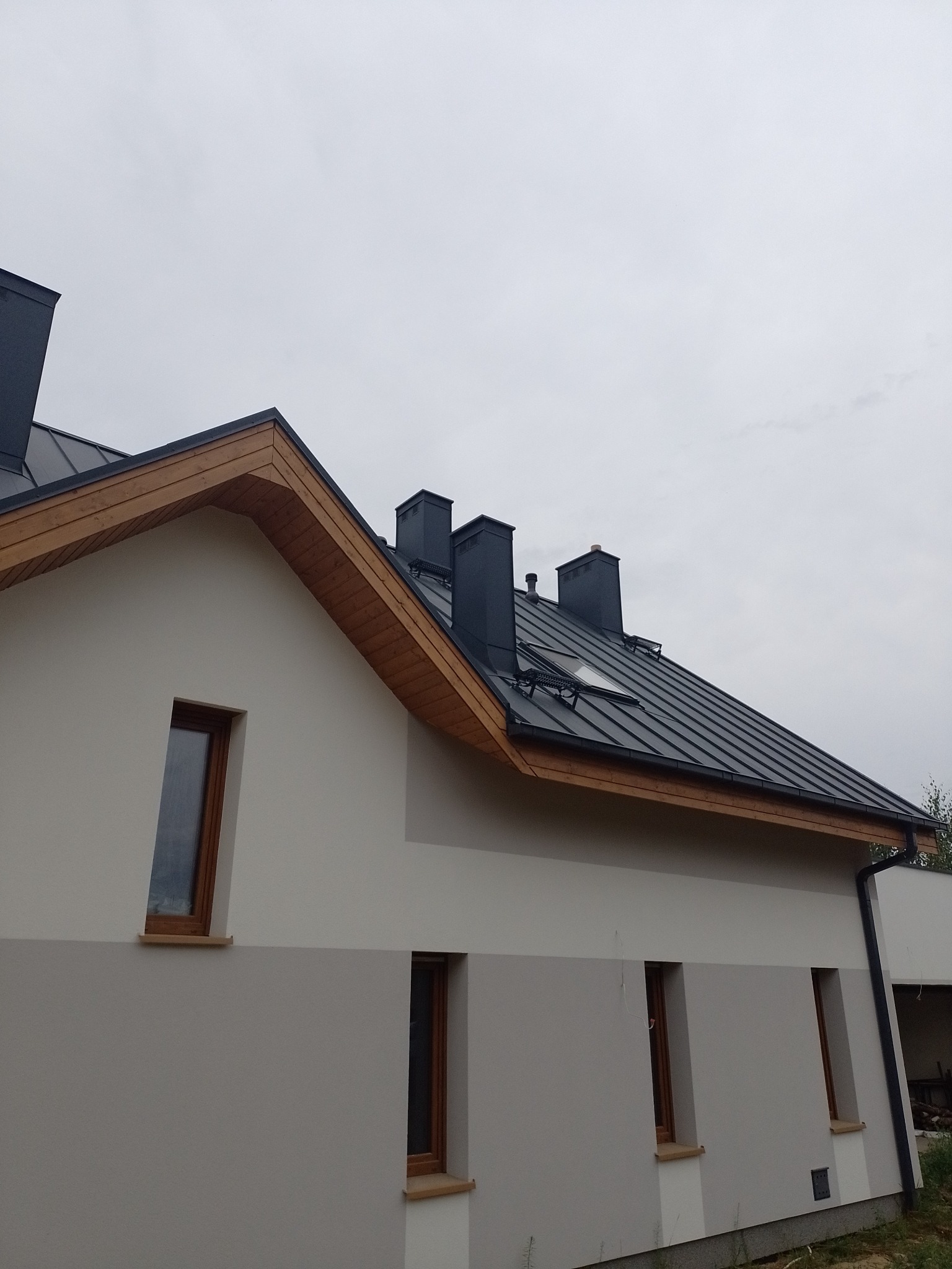 Nowoczesny dom z grafitowym dachem z blachy na rąbek stojący, drewnianą podbitką i trzema kominami, widok z boku.