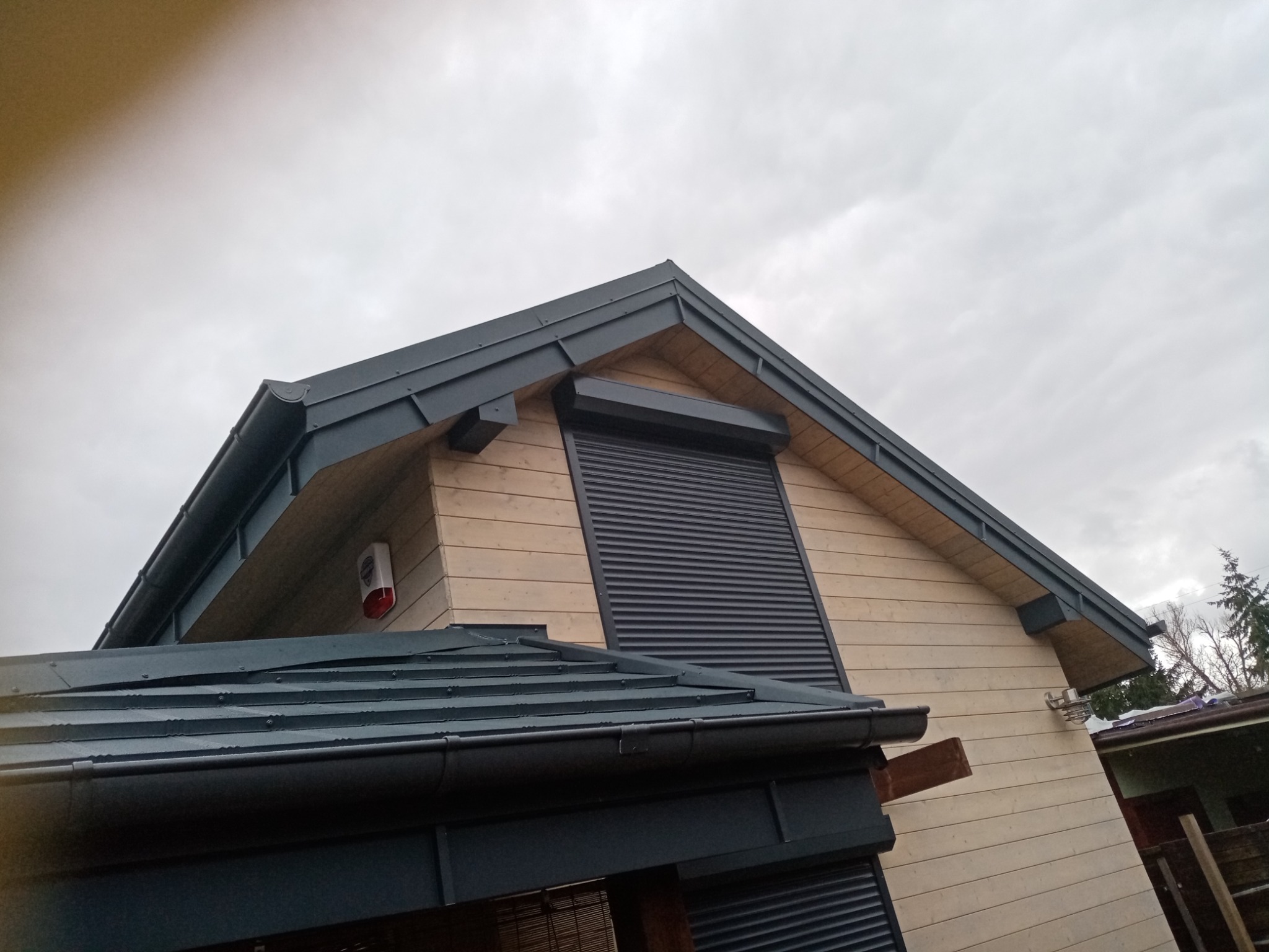 Fragment nowoczesnego domu z drewnianą elewacją, ciemnoszarym dachem z blachy i roletami zewnętrznymi, widoczny z dołu pod kątem, pochmurne niebo w tle.