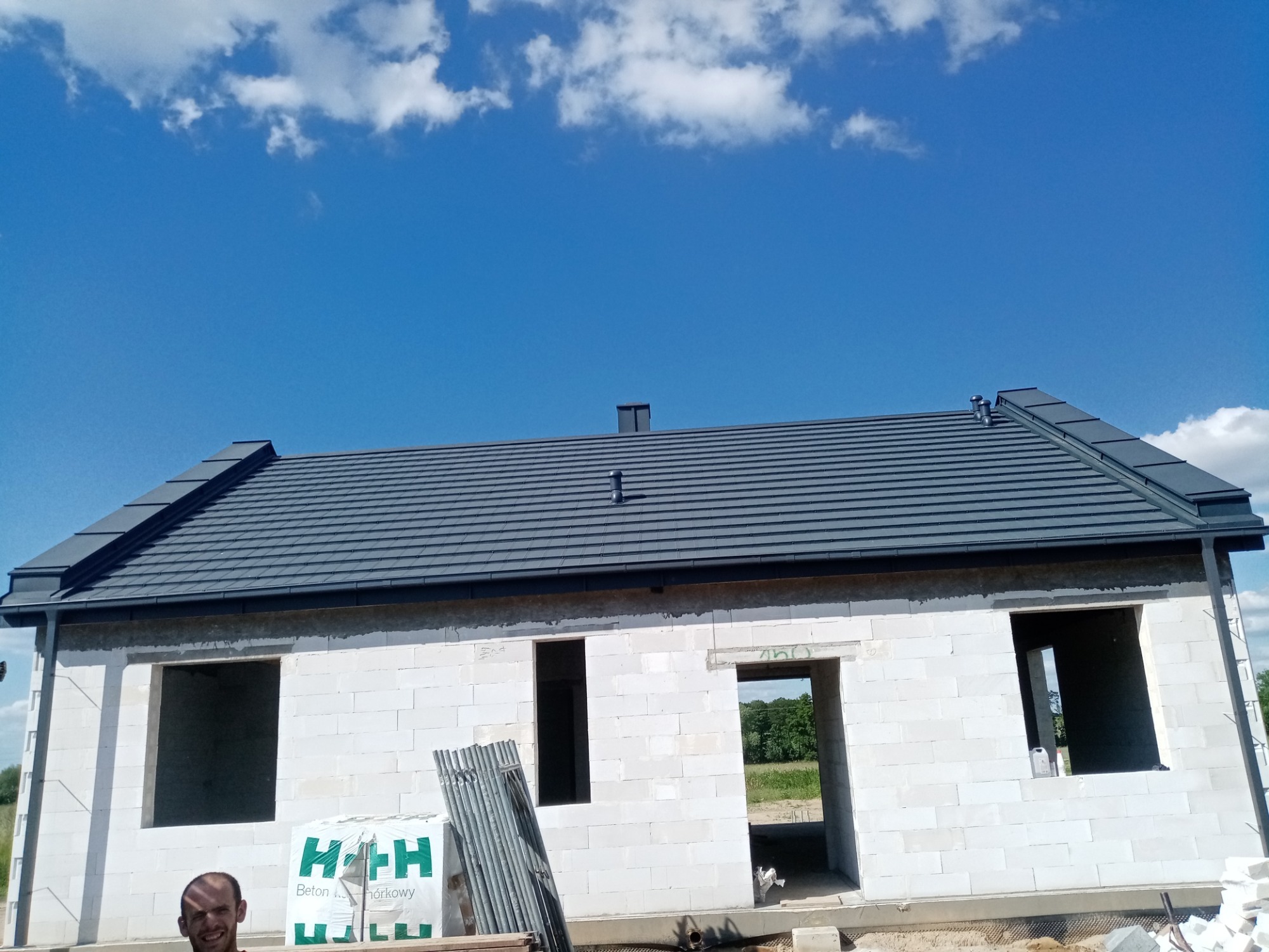 Surowy stan domu z białego bloczka z zamontowanym szarym dachem pod błękitnym niebem z chmurami, widoczne materiały budowlane i uśmiechnięta osoba w rogu kadru.