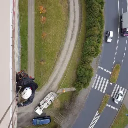 Pracownik wysokościowy w kasku i uprzęży, zawieszony na linie, wykonuje prace na elewacji wysokiego budynku, widok z góry, obok zaparkowany podnośnik koszowy, w tle chodnik, trawnik i ulica...