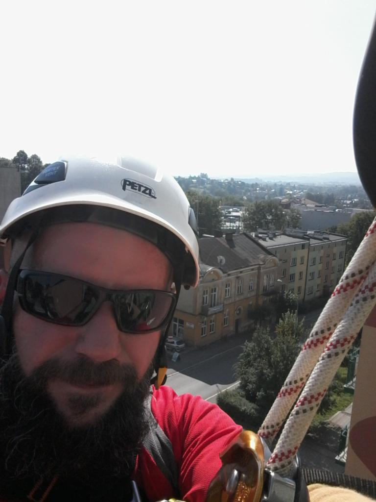 Mężczyzna z brodą w kasku Petzl i okularach przeciwsłonecznych, widok z wysokości na budynki miejskie i linę zabezpieczającą.
