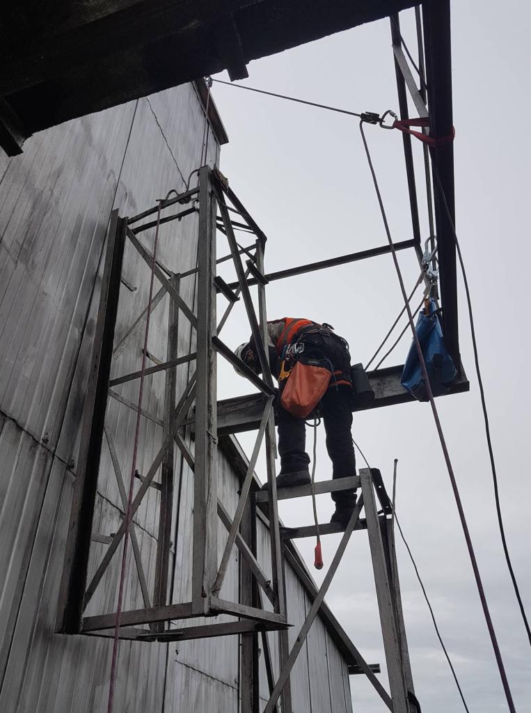 Pracownik w pomarańczowej kamizelce i kasku, zabezpieczony linami, na metalowej konstrukcji przy ścianie budynku z blachy trapezowej, wykonujący prace na wysokości.