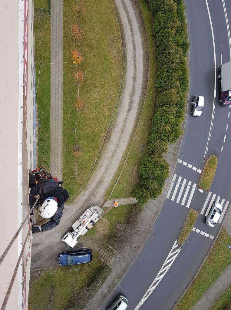 Pracownik wysokościowy w kasku i uprzęży, zawieszony na linie, wykonuje prace na elewacji wysokiego budynku, widok z góry, obok zaparkowany podnośnik koszowy, w tle chodnik, trawnik i ulica...