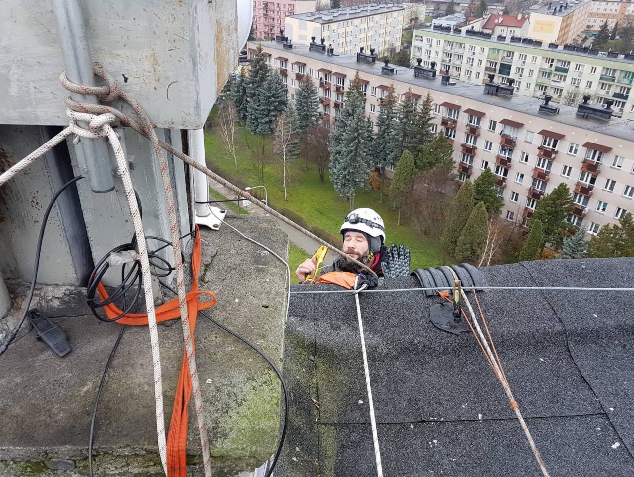 Pracownik w kasku, zabezpieczony linami, na dachu bloku mieszkalnego z widokiem na osiedle, podczas pracy na wysokości.