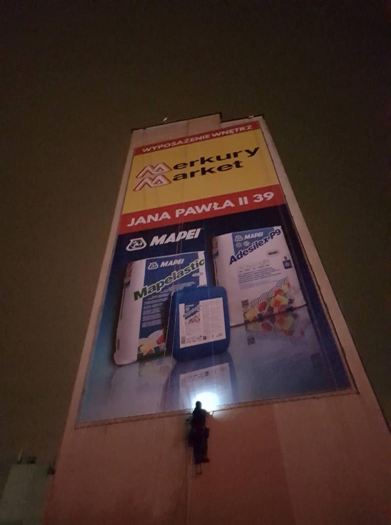 Pracownik na wysokościach zabezpieczony linami, wykonuje prace na ścianie budynku z dużym banerem reklamowym Merkury Market, na którym widoczne są produkty firmy Mapei.