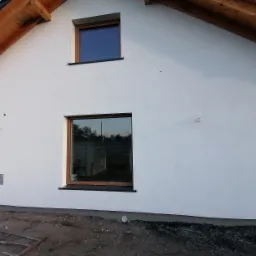 Biała elewacja domu jednorodzinnego z dwoma oknami o drewnianych ramach, widoczne ślady po montażu i niezakończone prace ziemne wokół budynku.