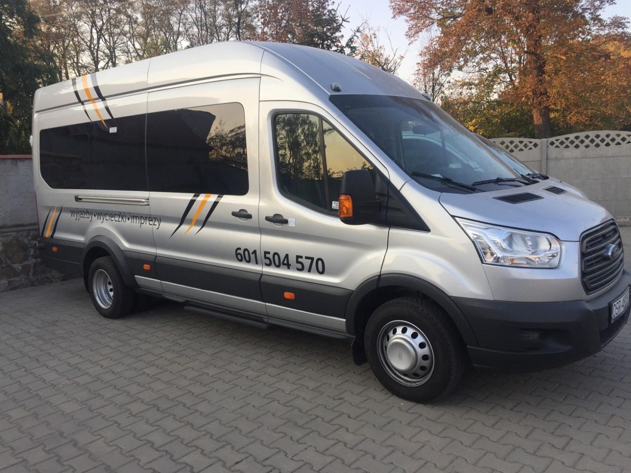 Srebrny bus Ford Transit z przyciemnianymi szybami i napisem 'wyjazdy wycieczki imprezy' oraz numerem telefonu na boku, zaparkowany na szarym bruku.