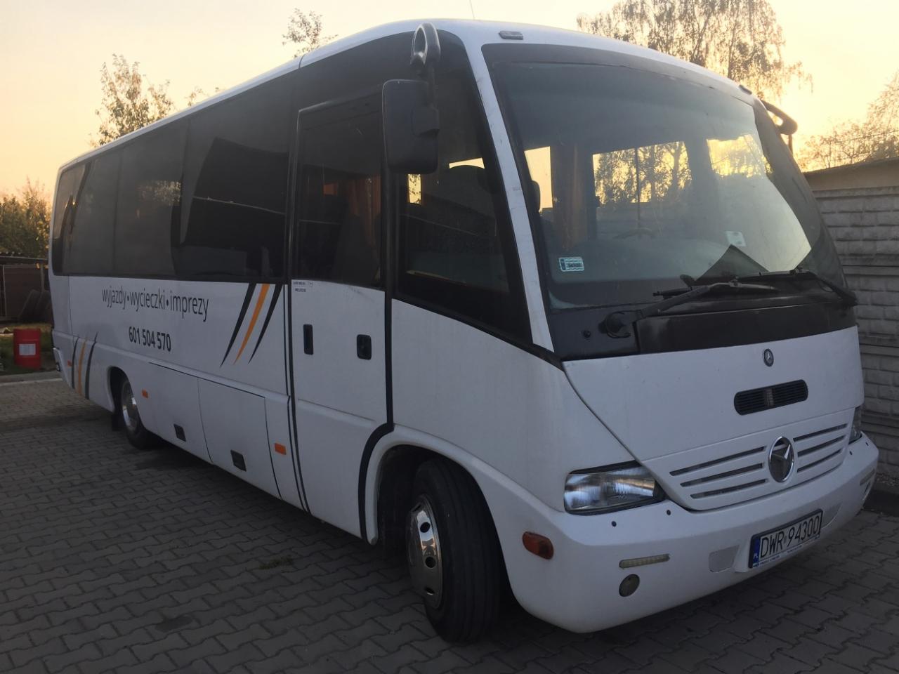 Biały bus turystyczny z numerem telefonu i napisem 'wyjazdy-wycieczki-imprezy' na boku, zaparkowany na brukowanym podjeździe o zmierzchu.