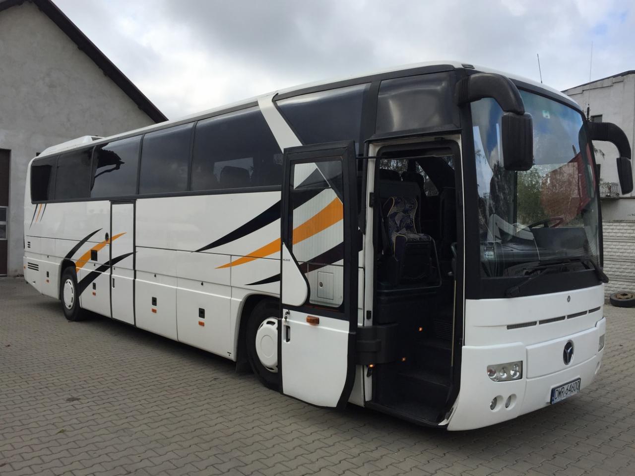 Biały autokar z otwartymi drzwiami, widoczne ozdobne detale w kolorze pomarańczowym i czarnym na karoserii, zaparkowany na brukowanym placu.