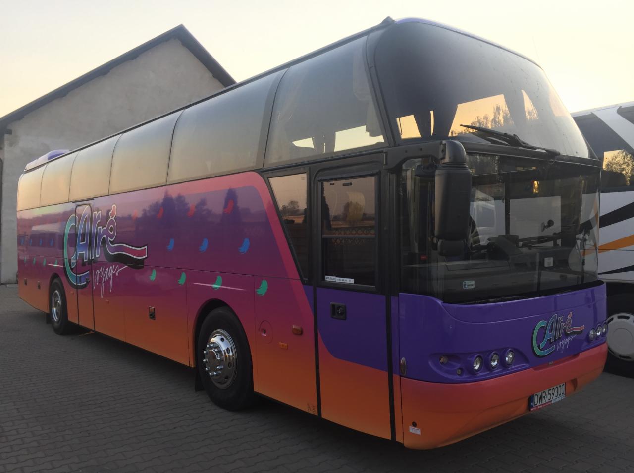 Autokar turystyczny z kolorowym malowaniem, widok z boku na tle budynku i innego pojazdu.