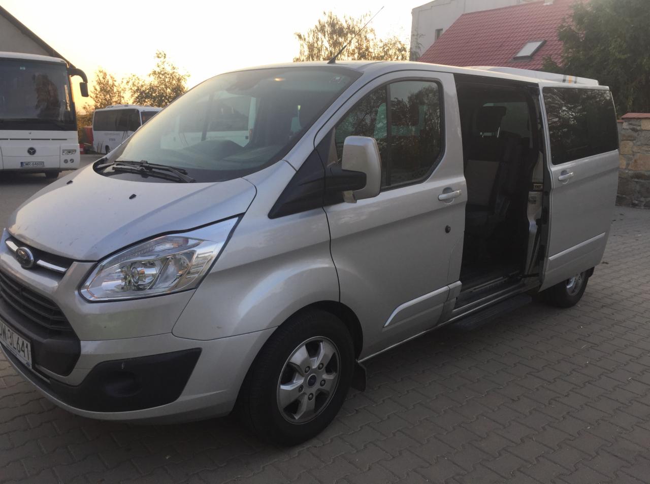 Srebrny van Ford Transit z otwartymi drzwiami przesuwnymi na brukowanym podjeździe, w tle autobus i budynek z czerwonym dachem.