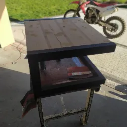 Stół warsztatowy z drewnianym blatem i czarną metalową ramą, umieszczony na metalowym stojaku, z motocyklem crossowym w tle.