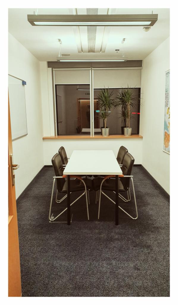 Klimatyczna sala konferencyjna z białym stołem, czarnymi krzesłami i szarym dywanem, oświetlona podłużną lampą sufitową, z widokiem na biuro przez okno z żaluzjami.