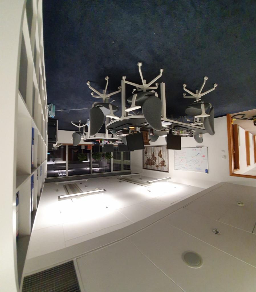 Nowoczesne biuro z białymi meblami, szarymi krzesłami obrotowymi i ciemnoniebieską wykładziną. Widoczne biurka z komputerami, regał z segregatorami i okno z widokiem na zieleń. Na ścianie mapa...