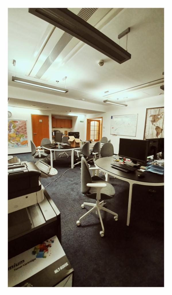 Biuro z ciemną wykładziną, okrągłymi stołami, szarymi krzesłami na kółkach, drukarką i kartonem na blacie, oświetlone lampami sufitowymi.