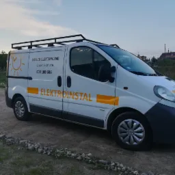 Biały samochód dostawczy z napisem 'Usługi Elektryczne Szymon Kelich' i numerem telefonu, zaparkowany na żwirowej drodze, z bagażnikiem dachowym.