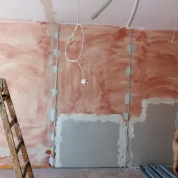 Ściana w trakcie przygotowywania do szpachlowania, widoczne profile metalowe, częściowo nałożona masa, drewniana drabina oparta o ścianę, fragment instalacji elektrycznej zwisający z sufitu.