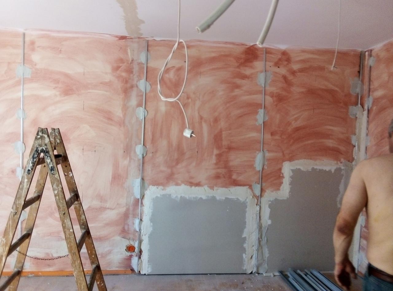 Ściana w trakcie przygotowywania do szpachlowania, widoczne profile metalowe, częściowo nałożona masa, drewniana drabina oparta o ścianę, fragment instalacji elektrycznej zwisający z sufitu.