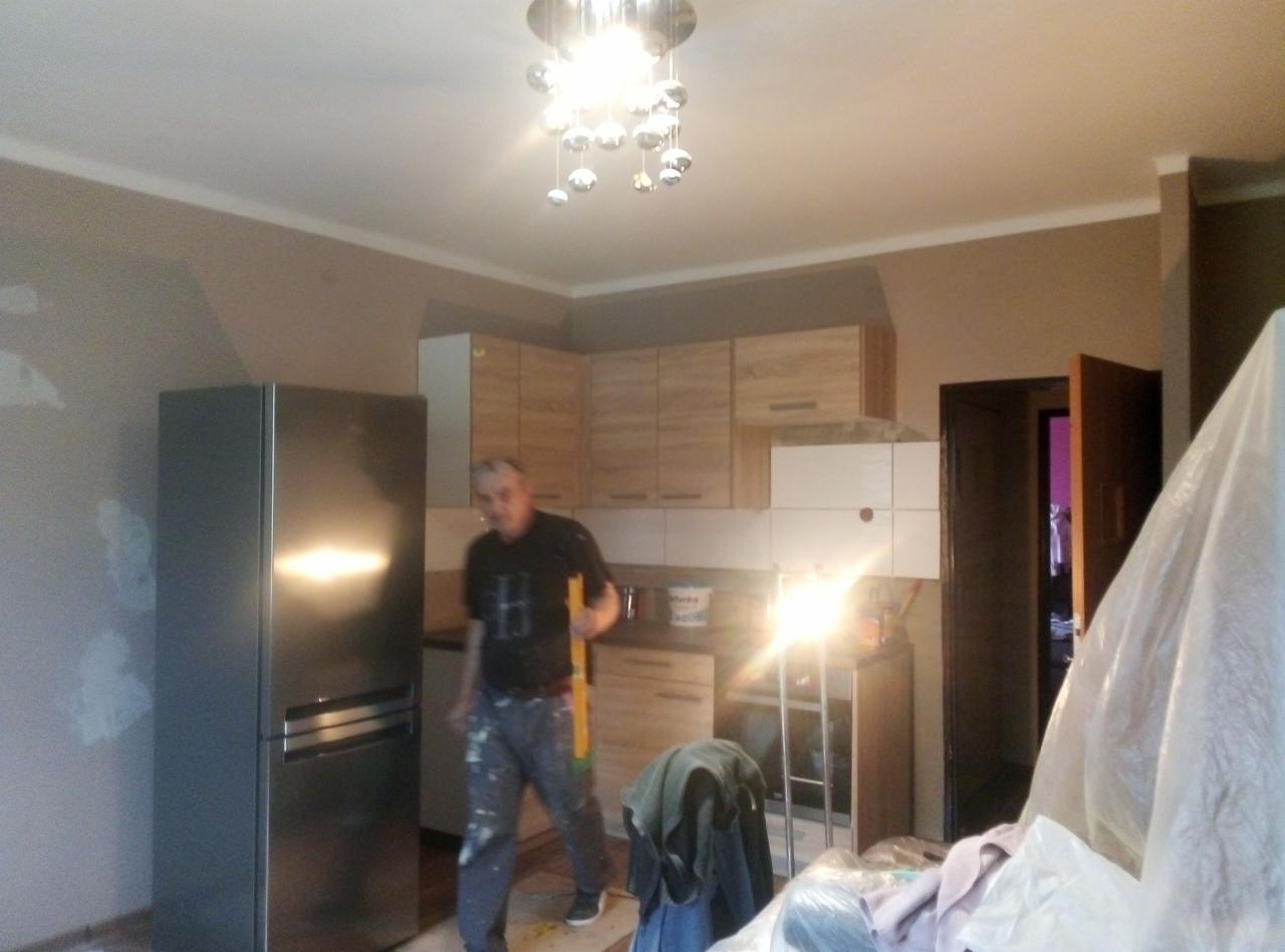Remont kuchni w trakcie: mężczyzna z poziomicą na tle lodówki, szafek i osłoniętych mebli. Ściana częściowo przygotowana do malowania.