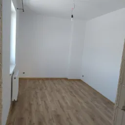 Pusty pokój po remoncie z panelami podłogowymi w odcieniach brązu, białymi ścianami, grzejnikiem pod oknem i wiszącą żarówką na kablu.