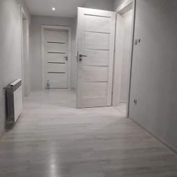 Siw-Bud - Jasne panele podłogowe w korytarzu z szarymi ścianami, białe drzwi wewnętrzne z poziomymi frezami, grzejnik na ścianie po lewej stronie.