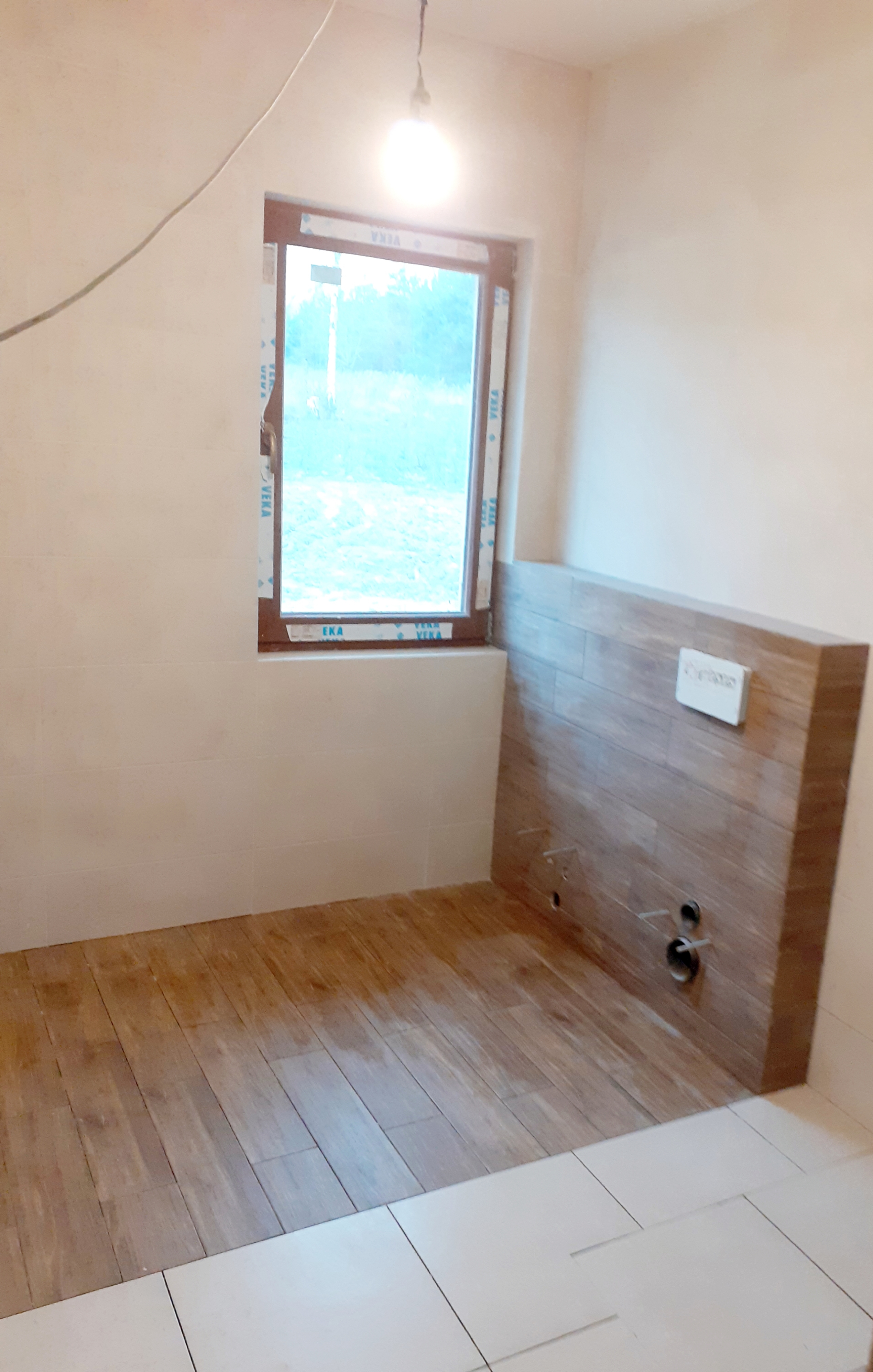 Wnętrze łazienki w trakcie remontu: ułożona drewnopodobna glazura na podłodze i obudowie stelaża WC, białe płytki na ścianach do połowy wysokości, okno z widokiem na zieleń.