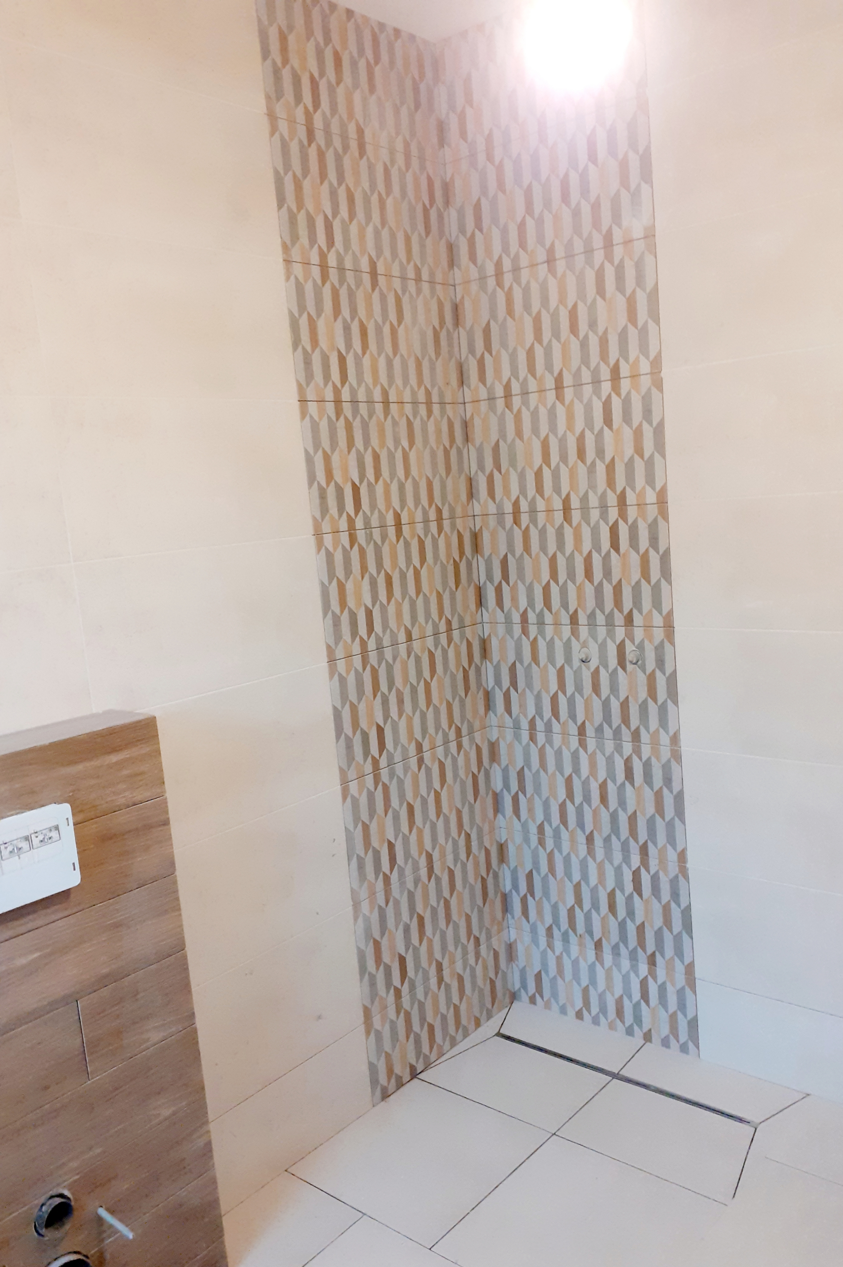 Wykończona ściana w łazience z dekoracyjnymi płytkami w geometryczny wzór, kremowe płytki na ścianach i podłodze, widoczny odpływ liniowy i zabudowa WC z drewnopodobną okleiną.