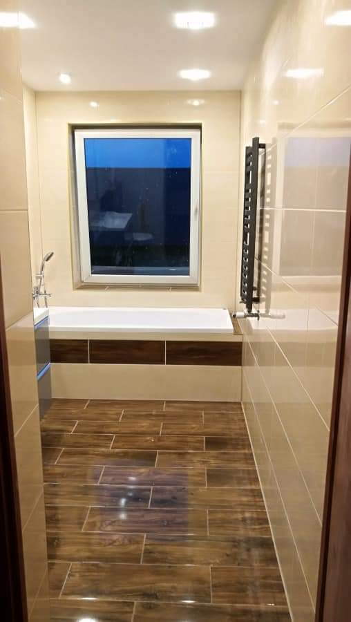 Wykończona łazienka z drewnopodobną podłogą z płytek, beżowymi płytkami na ścianach, białą wanną obudowaną panelami w kolorze drewna i drabinkowym grzejnikiem.