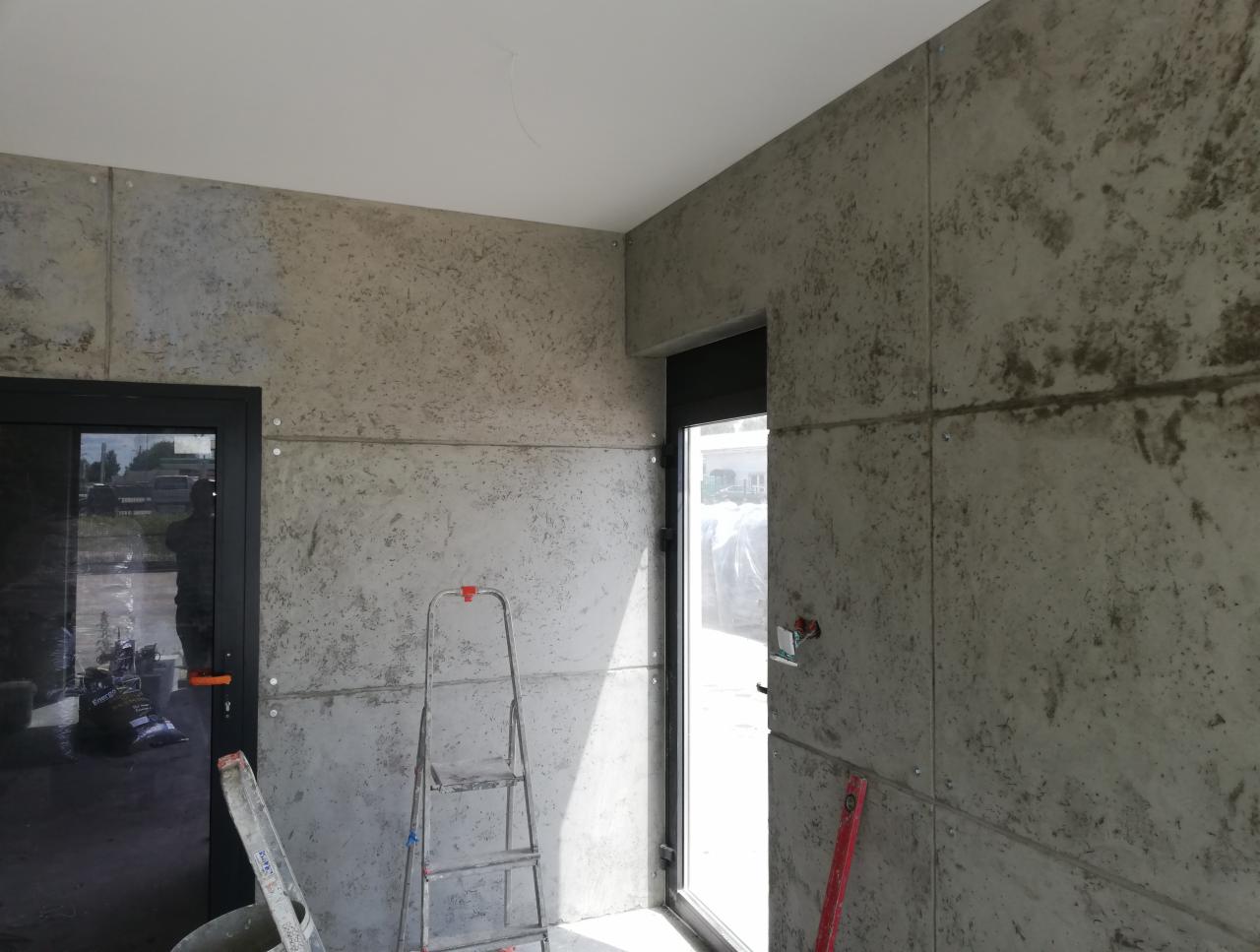 Wnętrze z szarymi płytami imitującymi beton na ścianach, widoczna drabina i drzwi. Surowy, nowoczesny styl wykończenia wnętrza.