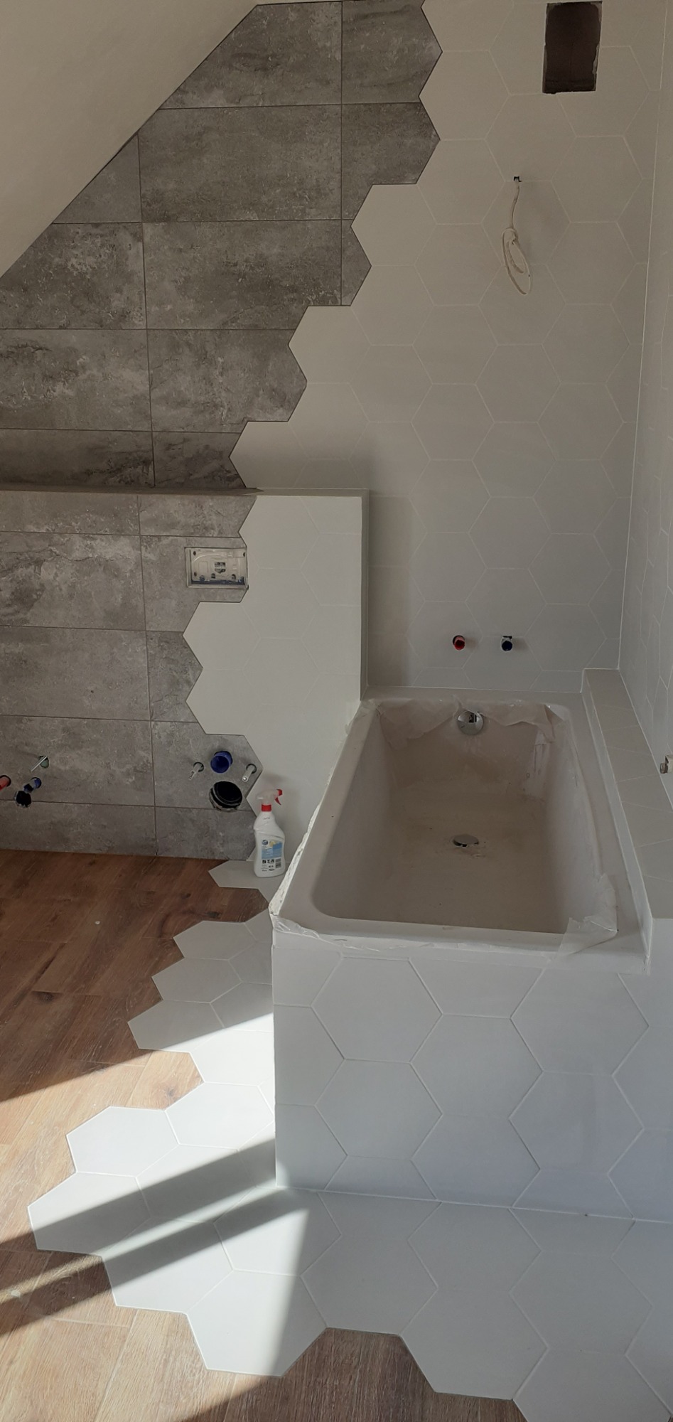 Łazienka w trakcie remontu: ściany częściowo wyłożone szarymi i białymi heksagonalnymi płytkami, wanna zabezpieczona folią, widoczne instalacje wodne i fragment podłogi z paneli.